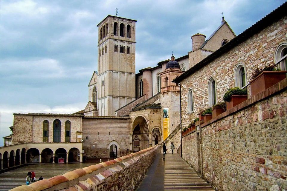 Una basílica histórica de piedra con un alto campanario se alza junto a una amplia rampa en un patio bajo un cielo nublado.
