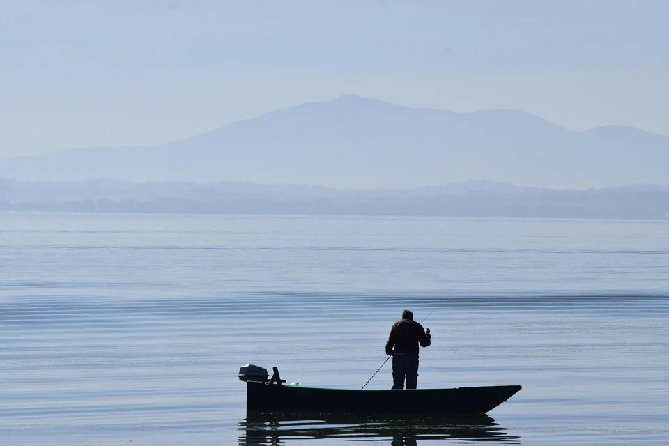 Una persona de pie en una pequeña lancha motora, pescando en aguas tranquilas con montañas brumosas en la distancia.