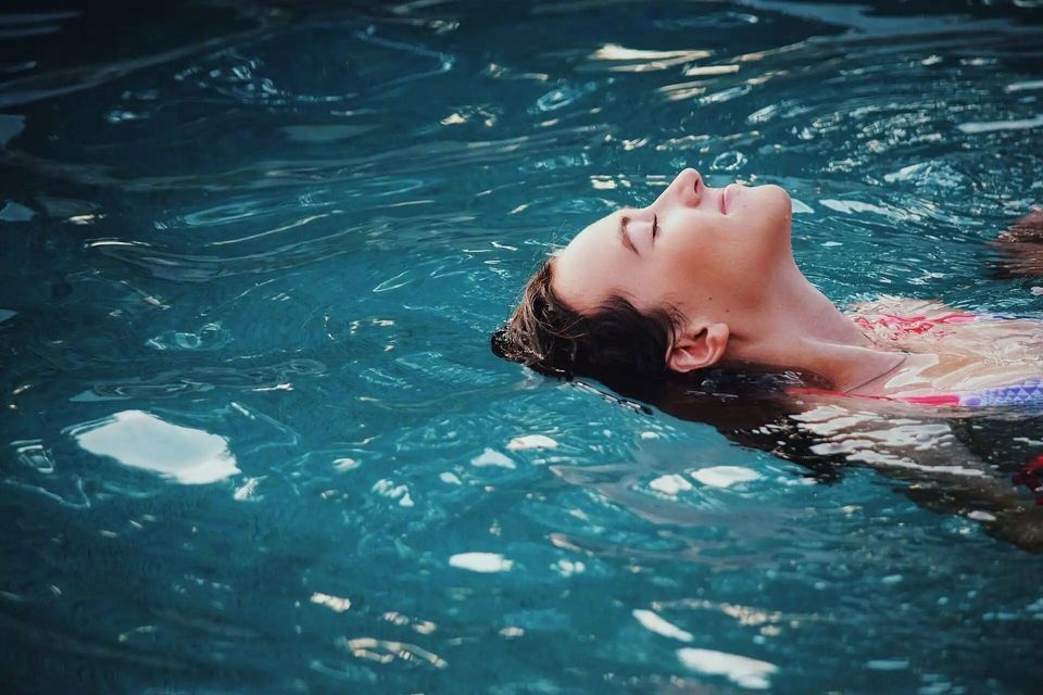 Una mujer flota de espaldas con los ojos cerrados en agua azul cristalina, con la cabeza y los hombros sobre la superficie.