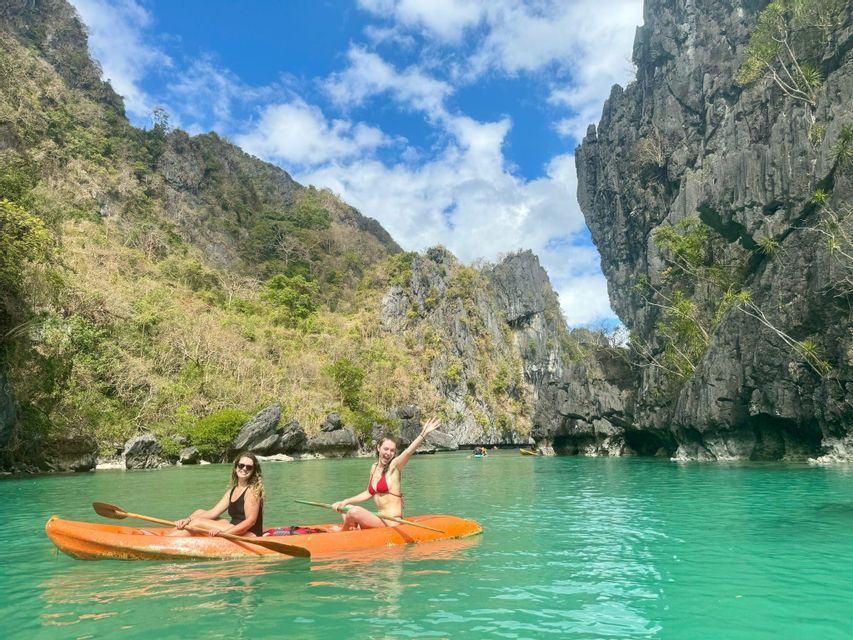 Dos mujeres en un viaje grupal de WeRoad reman en un kayak naranja sobre agua turquesa entre grandes acantilados rocosos bajo un cielo azul nublado.