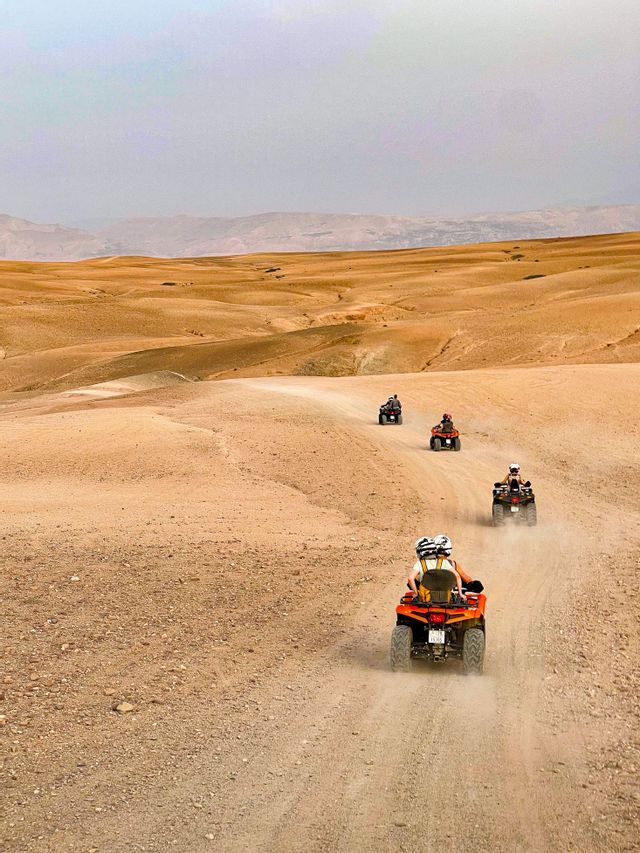 A WeRoad group trip riding quad bikes in a line on a dusty trail through a vast desert landscape.