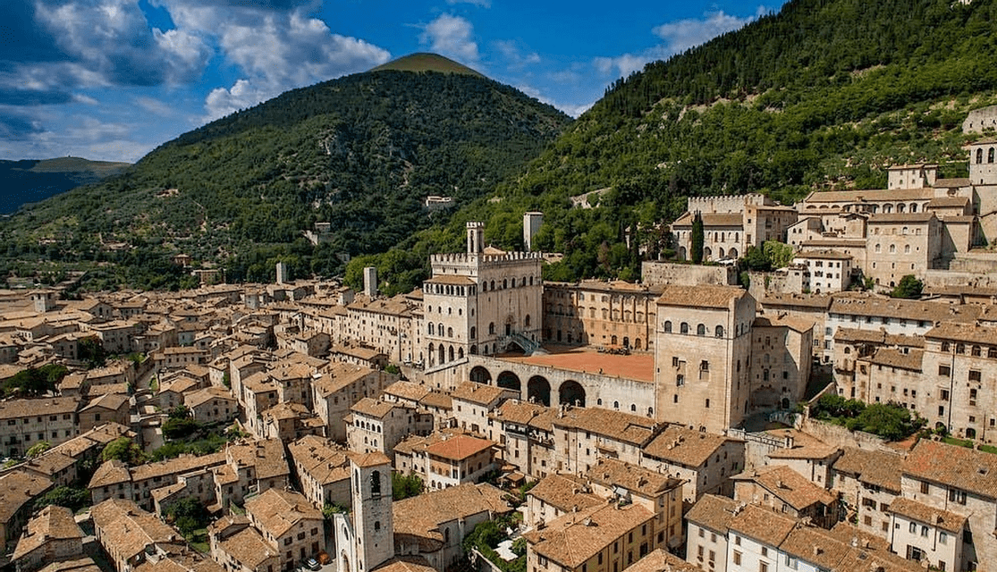 Una vista aerea di una città storica con tetti in terracotta, costruita alla base di una montagna lussureggiante e verde sotto un cielo azzurro.