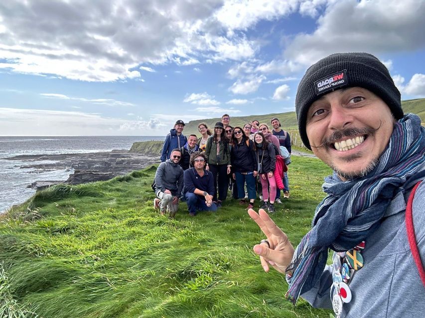 Un hombre se hace un selfi con un viaje en grupo de WeRoad en un acantilado cubierto de hierba con vistas a una costa rocosa bajo un cielo nublado.