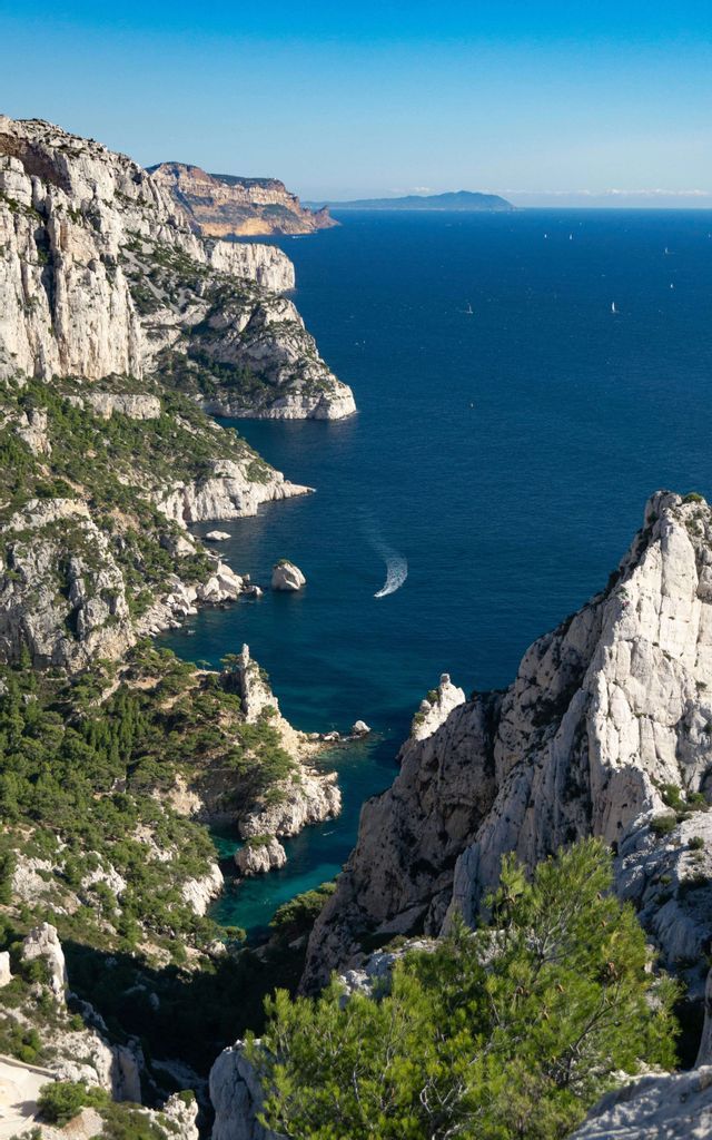 Vista aérea de una costa con acantilados blancos y empinados, árboles verdes y un mar azul profundo con un pequeño barco navegando.
