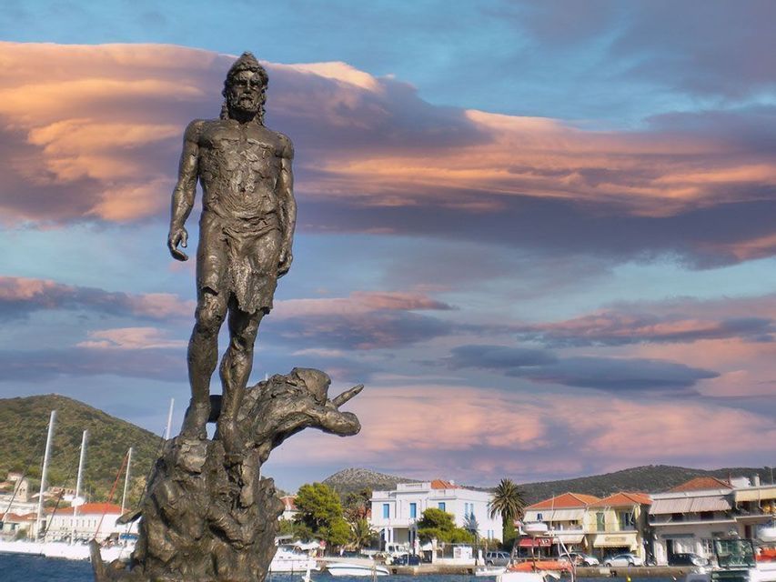 Una estatua de bronce de un hombre se alza sobre un pedestal con vistas a un pueblo costero y su puerto al atardecer.