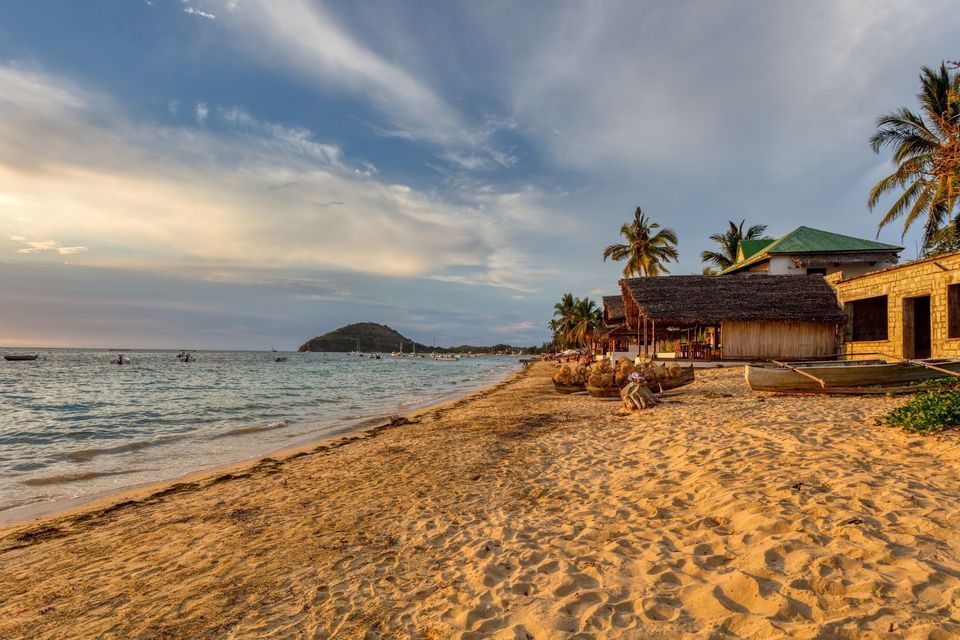 Ein Sandstrand mit Strohdachhütten und Booten am Ufer während eines goldenen Sonnenuntergangs.