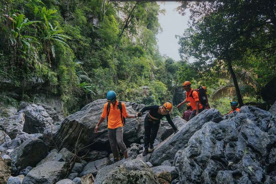 Un gruppo WeRoad in viaggio, con caschi e magliette arancioni brillanti, attraversa con cautela grandi massi in un lussureggiante canyon nella giungla.