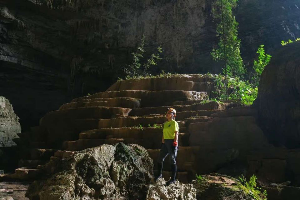Una persona con un casco si trova all'interno di una grande grotta illuminata dal sole, osservando gigantesche formazioni rocciose a terrazze.