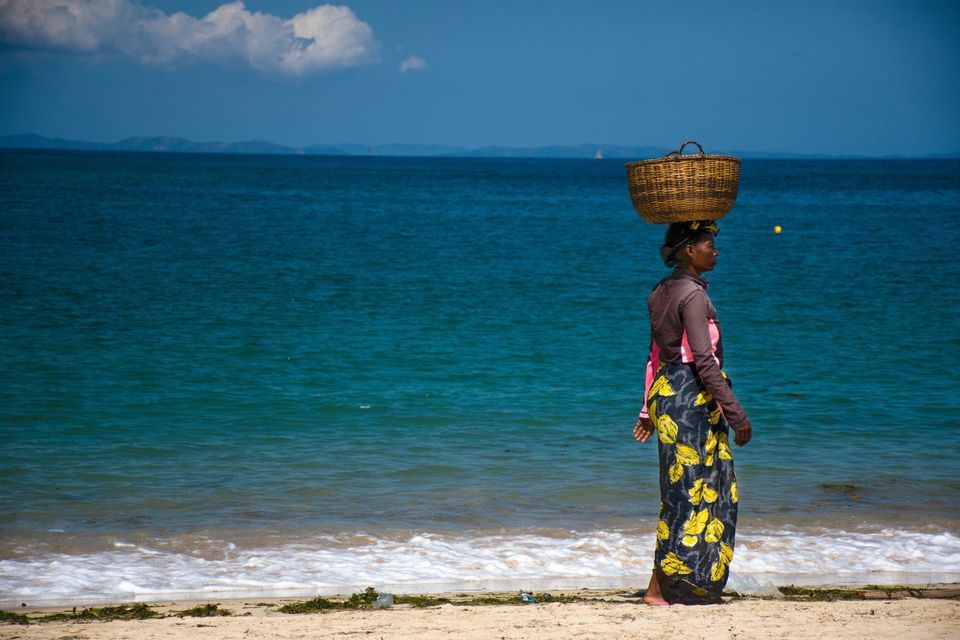Eine Frau geht an einem Sandstrand am blauen Ozean entlang und balanciert einen großen Weidenkorb auf ihrem Kopf unter klarem Himmel.