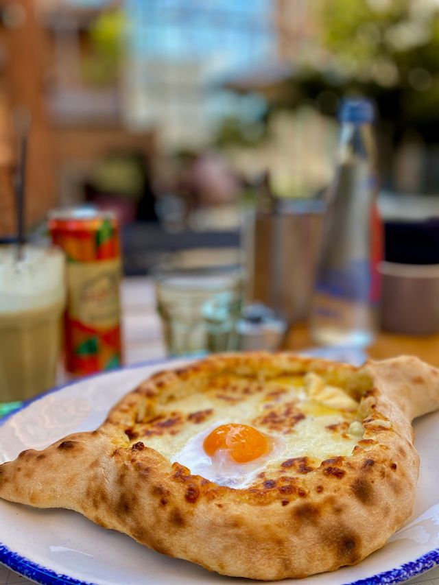 Un Khachapuri Adjaruli, un pane a forma di barca con formaggio e un uovo, è adagiato su un piatto su un tavolo.