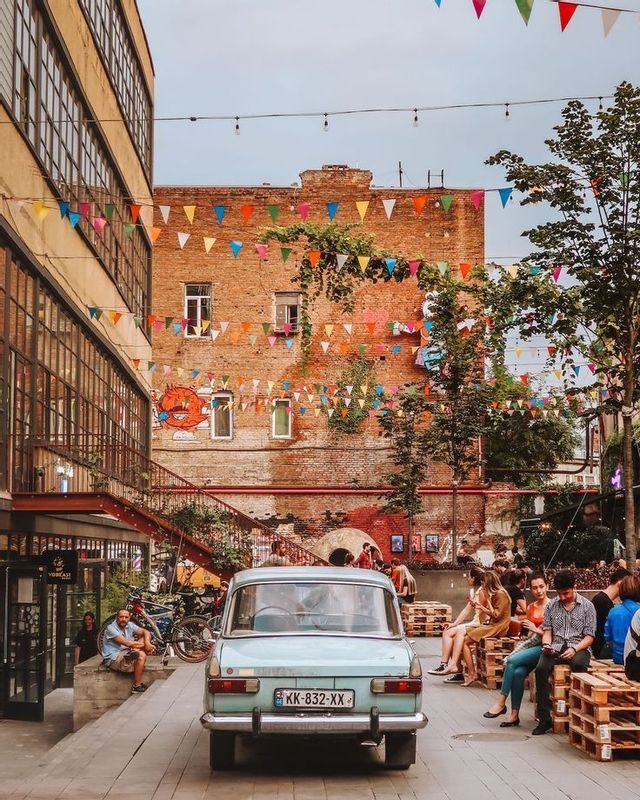 Un'auto d'epoca blu parcheggiata in un cortile urbano festoso decorato con bandierine colorate mentre le persone si rilassano su sedute di pallet.