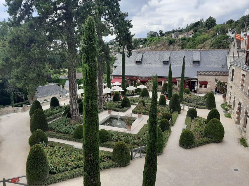 Vue plongeante sur un jardin formel avec topiaires, allées de gravier et une fontaine centrale, à côté d'un bâtiment doté d'un café en plein air.