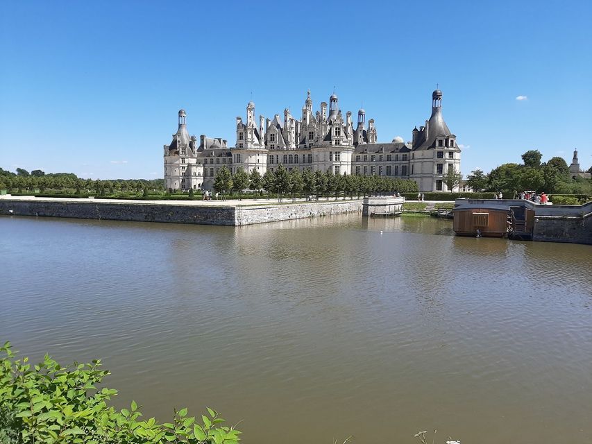 Un grand château de pierre blanche, orné de nombreuses tourelles, se dresse derrière de larges douves, sous un ciel bleu dégagé.