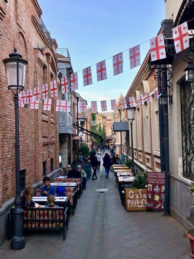 Una strada stretta con edifici in mattoni è fiancheggiata da tavolini di caffè all'aperto e decorata con festoni di bandiere.