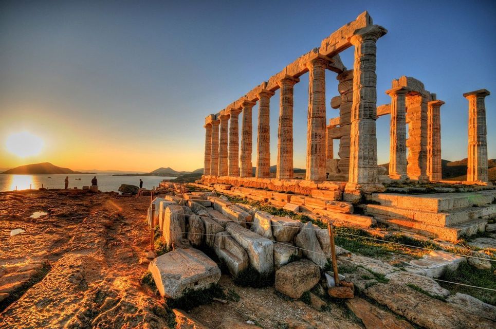 Le rovine di un antico tempio con grandi colonne si ergono su una costa rocciosa al tramonto sul mare.