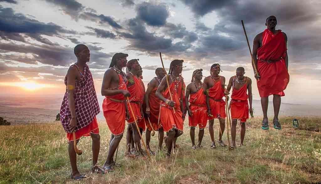 Un grupo de hombres masái con vestimenta roja tradicional en una colina de hierba, con uno realizando un salto alto al atardecer.