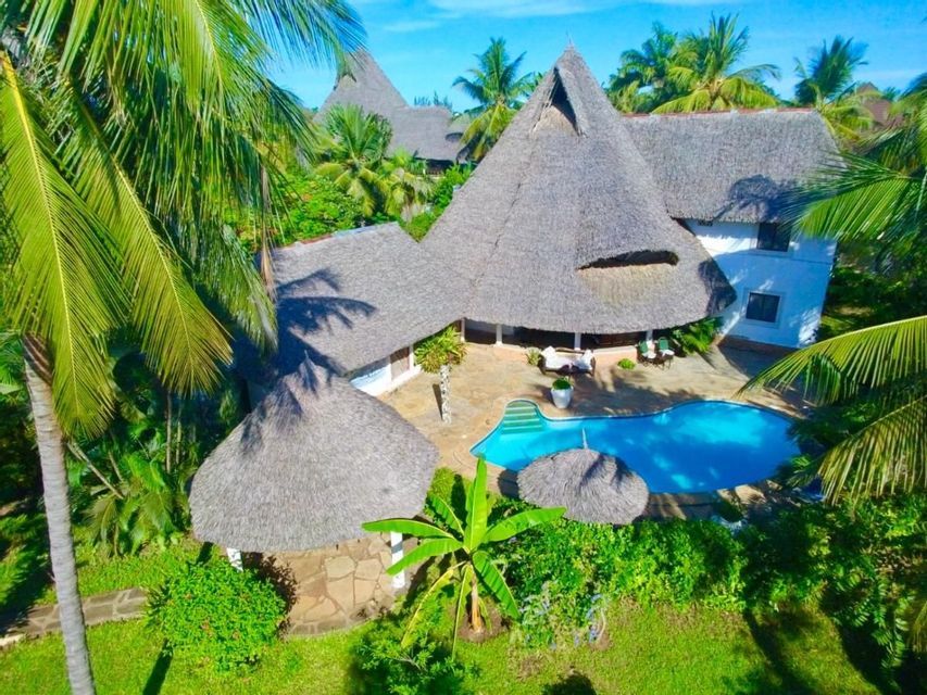 Una vista aérea de una villa tropical con techos de paja y una piscina, rodeada de exuberantes palmeras verdes.
