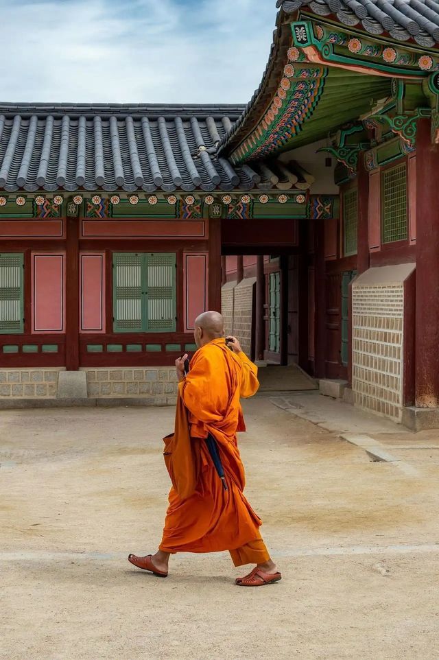 A Buddhist monk in bright orange robes walks across a sandy courtyard in front of a traditional temple.
