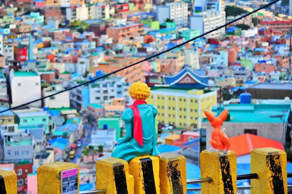 Figurines of The Little Prince and a fox sit on yellow bollards, overlooking a dense, colorful city below.