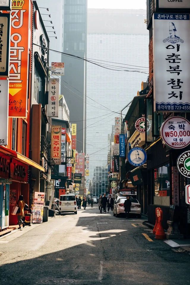 Pedestrians walk down a sunlit, narrow street lined with buildings covered in colorful Korean signs, with a modern skyscraper in the background.