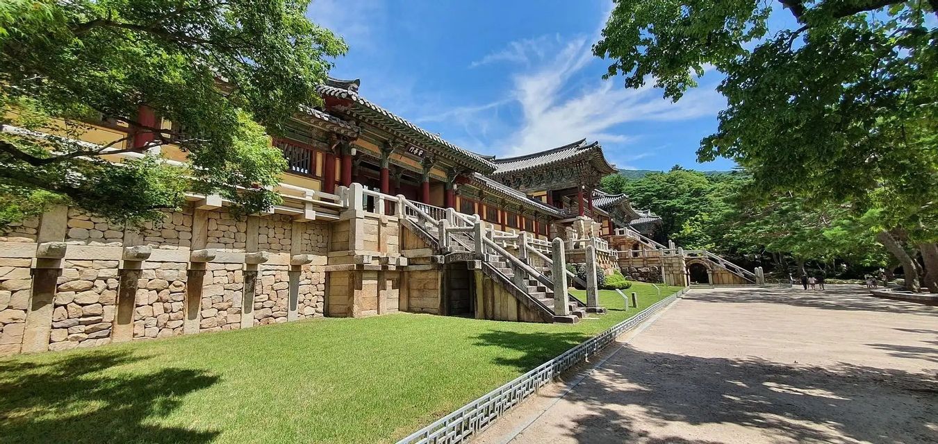A traditional Korean temple with a stone foundation and stairs, set against a green lawn and trees under a blue sky.