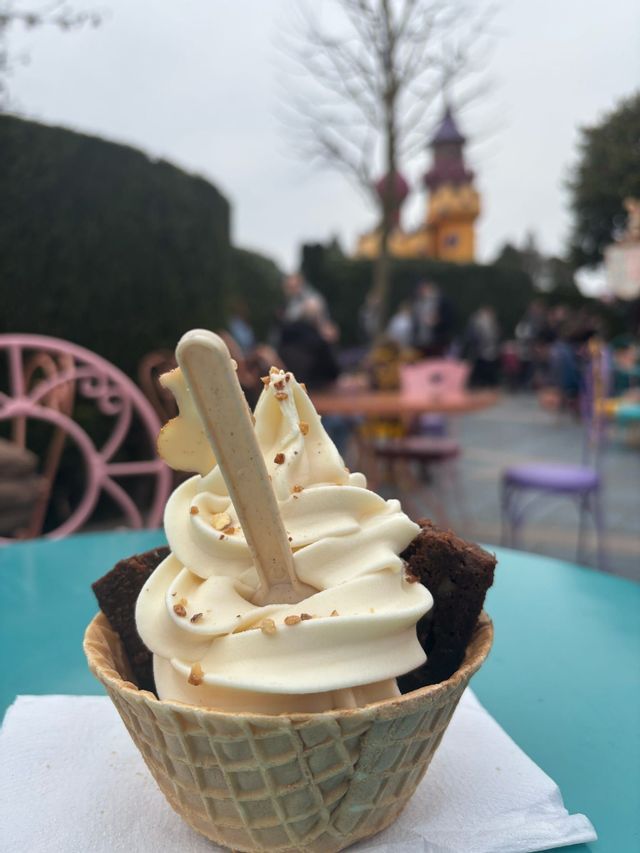 Un primo piano di un gelato soft in una cialda con pezzi di brownie e noci, su un tavolo color verde acqua all'aperto.