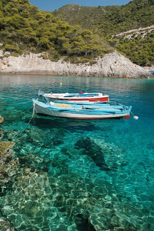 Tres pequeñas barcas, una azul y una roja, flotan en agua turquesa transparente junto a una ladera rocosa y arbolada.