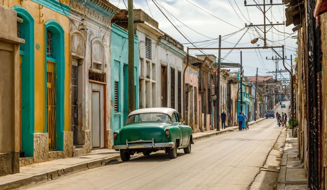 Un'auto d'epoca verde parcheggiata su una strada fiancheggiata da edifici colorati e logori sotto una ragnatela di cavi elettrici aerei.