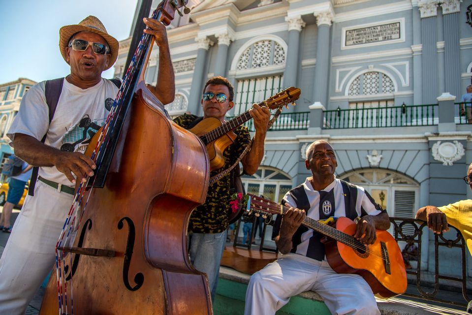 Un gruppo di musicisti di strada suona un contrabbasso e due chitarre davanti a un grande edificio grigio in una giornata di sole.
