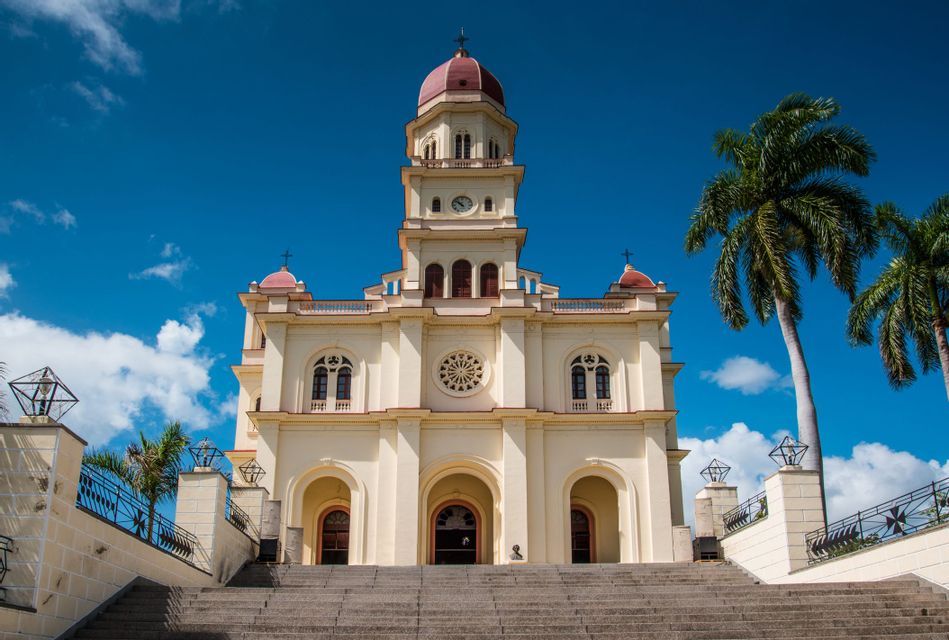 Una grande chiesa color crema con una cupola rossa si erge in cima a un'ampia scalinata in pietra, incorniciata da palme sotto un cielo blu.