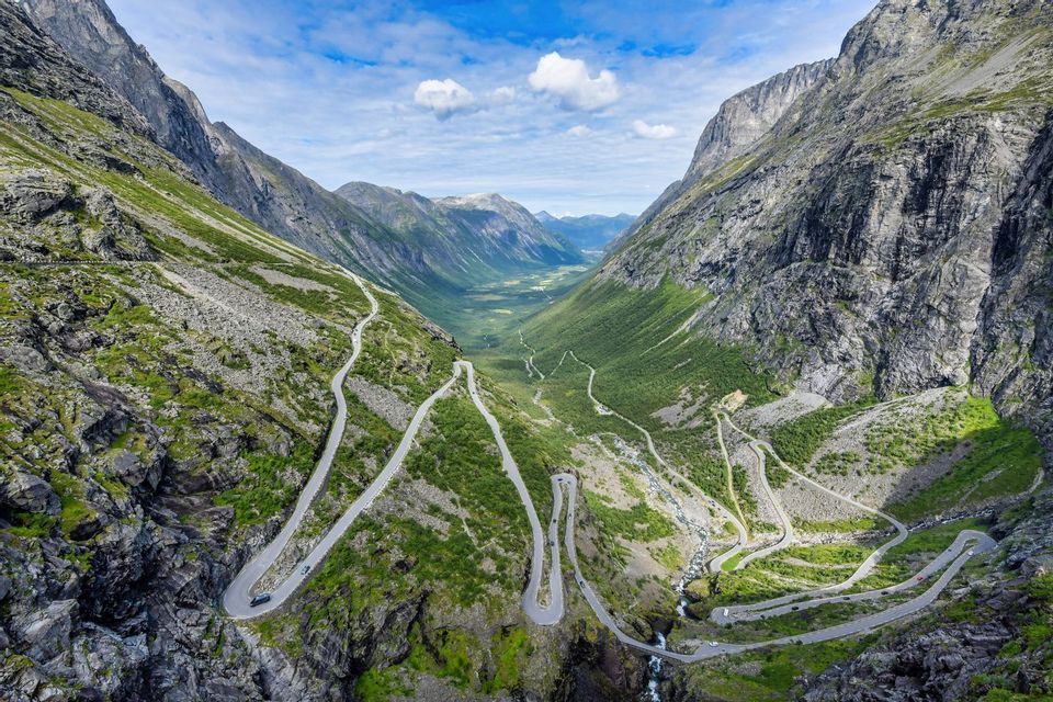 Una vista aerea di una strada tortuosa con tornanti che scende attraverso una ripida e verde valle di montagna sotto un cielo parzialmente nuvoloso.