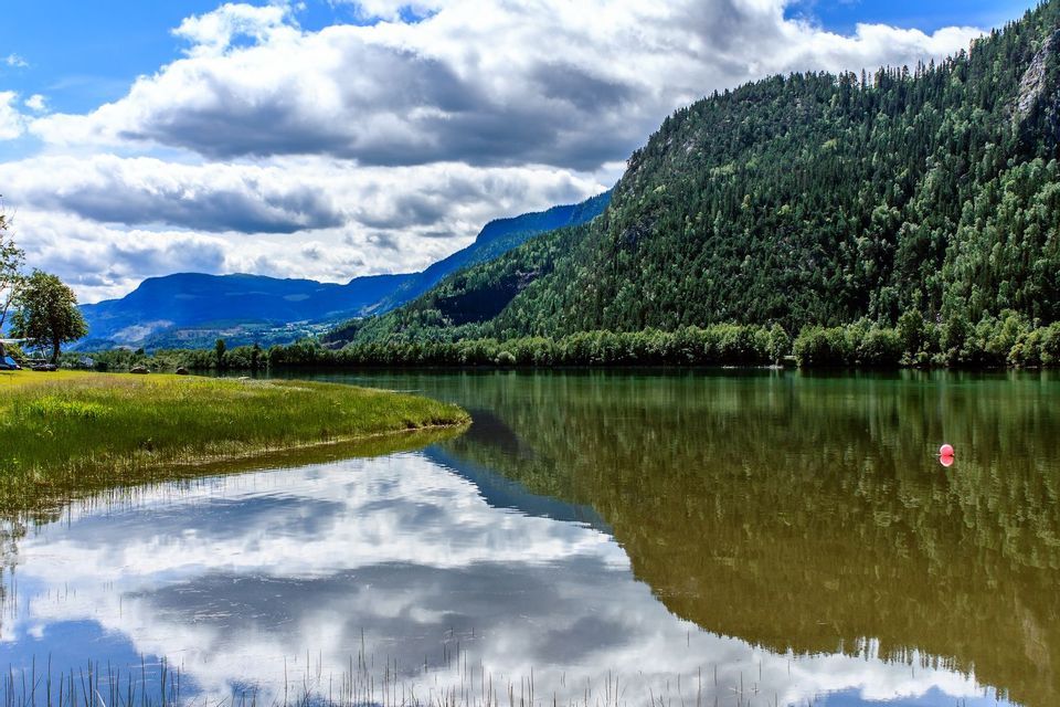 La superficie di un lago calmo riflette montagne verdi e alberate e un cielo parzialmente nuvoloso.
