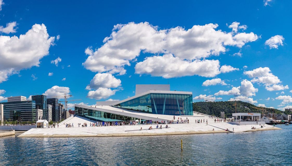 Un edificio moderno, con tetto bianco spiovente e pareti in vetro, si affaccia sull'acqua sotto un cielo azzurro intenso con nuvole soffici.