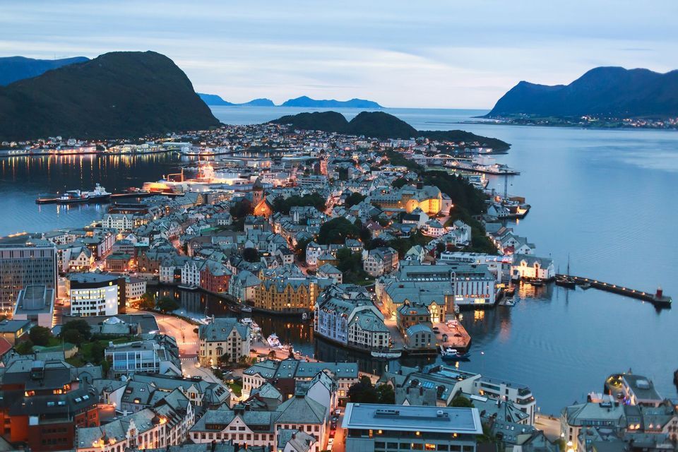Una vista aerea di una città costiera al tramonto, con le sue luci che si riflettono sull'acqua e grandi montagne sullo sfondo.