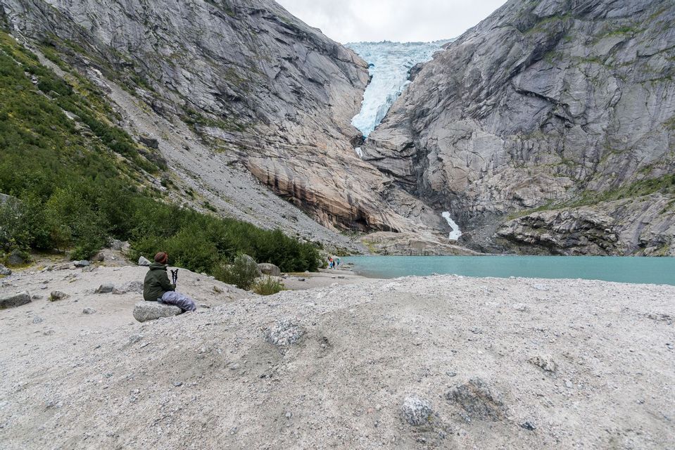 Una persona con bastoncini da trekking riposa su una roccia che si affaccia su un lago glaciale turchese e un imponente ghiacciaio di montagna.