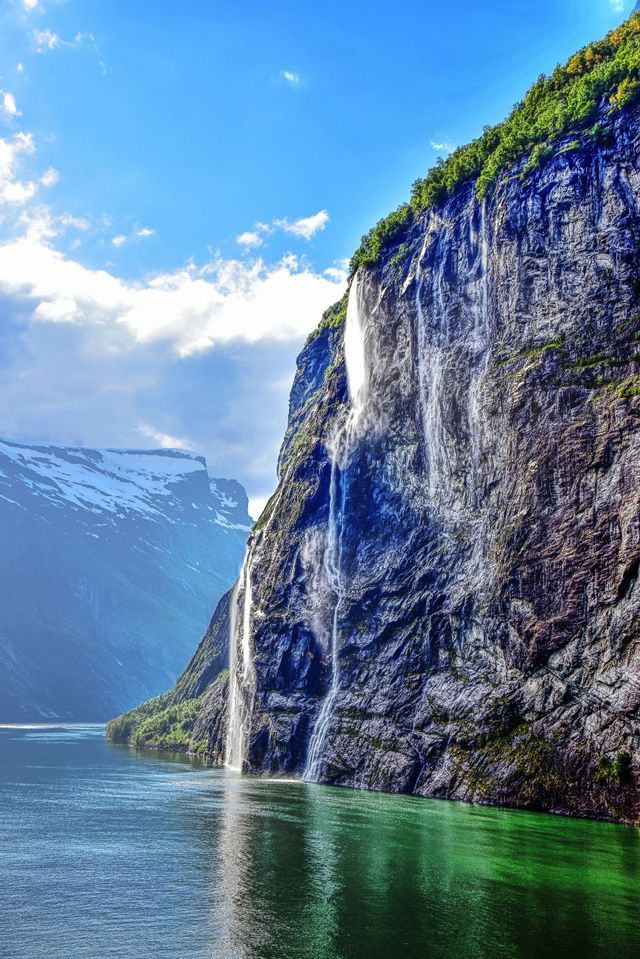 Cascate precipitano da un'alta scogliera verdeggiante in un fiordo, con montagne innevate sotto un cielo azzurro.