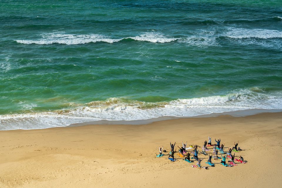 A WeRoad group trip practicing yoga on colorful mats on a sandy beach beside the turquoise ocean.