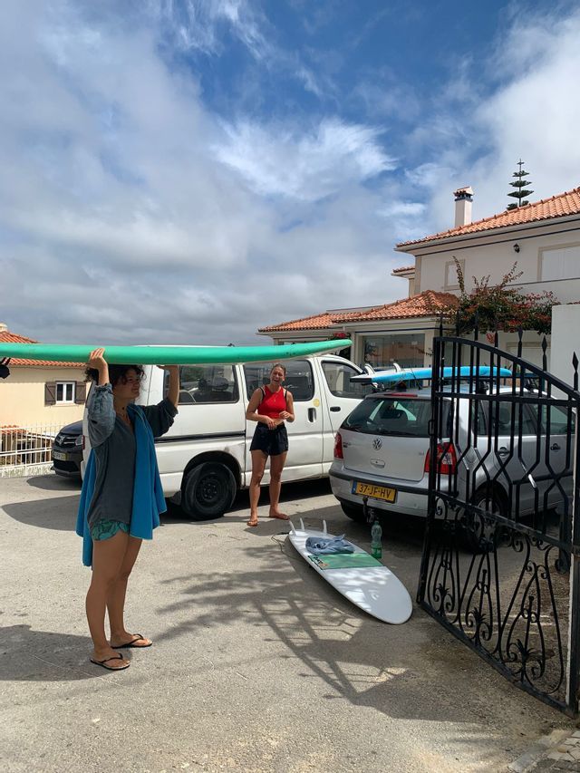Una donna in viaggio di gruppo WeRoad porta una tavola da surf verde in testa, mentre l'amica le sorride, in un vialetto con auto.