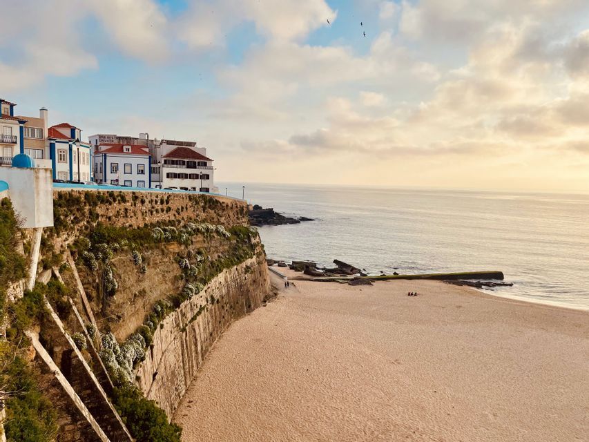 White buildings stand on a fortified cliff overlooking a wide sandy beach and the ocean under a cloudy sky.