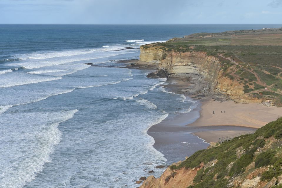 A high-angle view of a coastline with waves rolling onto a sandy beach next to tall, layered cliffs.