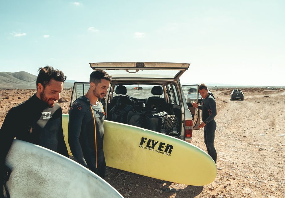 A WeRoad group trip of three men in wetsuits holding surfboards next to an SUV with its trunk open in an arid landscape.