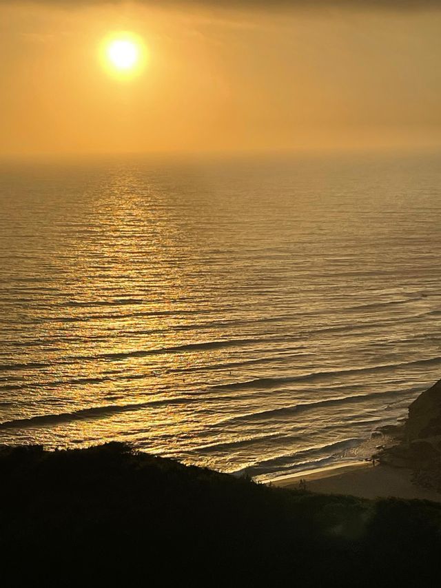 The sun sets over a hazy ocean, casting a golden reflection on the water as waves roll towards a silhouetted shore.