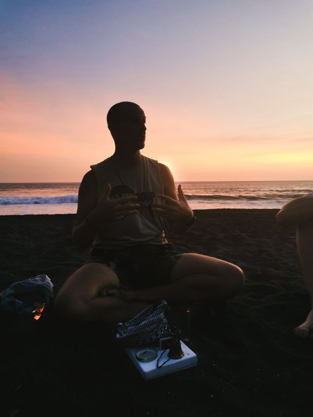 Eine Person einer WeRoad-Gruppenreise sitzt als Silhouette im Schneidersitz an einem dunklen Sandstrand und unterhält sich mit anderen, während die Sonne über dem Meer untergeht.