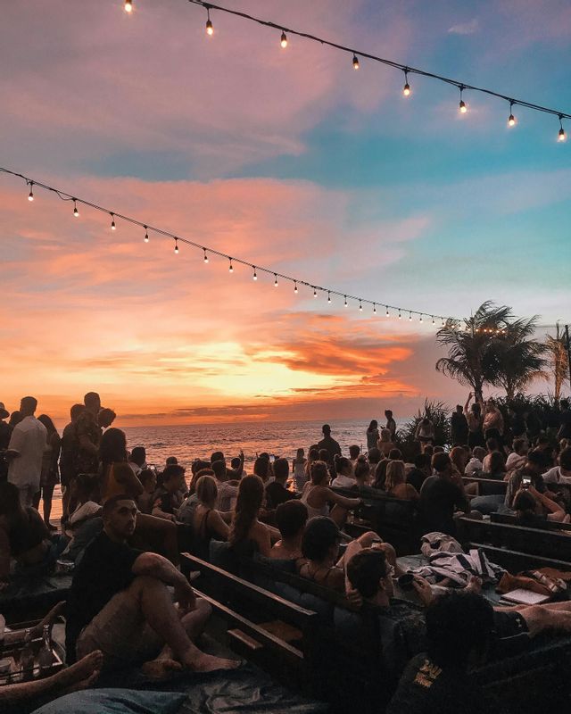 Eine WeRoad-Gruppe sitzt auf Strandliegen und beobachtet den Sonnenuntergang über dem Meer, mit Lichterketten über sich.