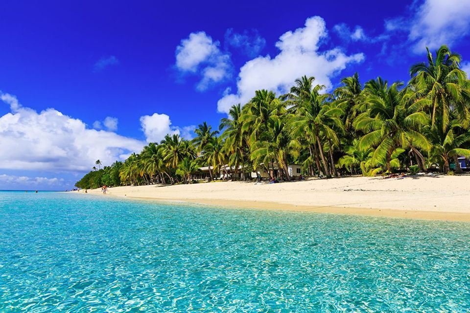 Una spiaggia tropicale con sabbia bianca e acqua cristallina turchese, fiancheggiata da palme sotto un cielo azzurro con nuvole.