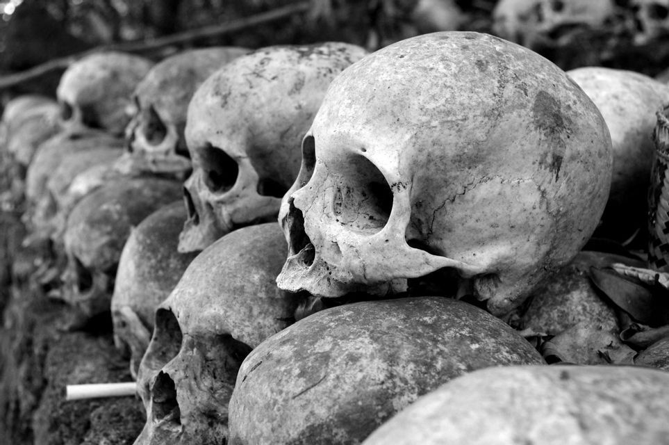 Schwarz-Weiß-Nahaufnahme von zahlreichen verwitterten menschlichen Schädeln, in Reihen gestapelt und von der Seite gesehen.