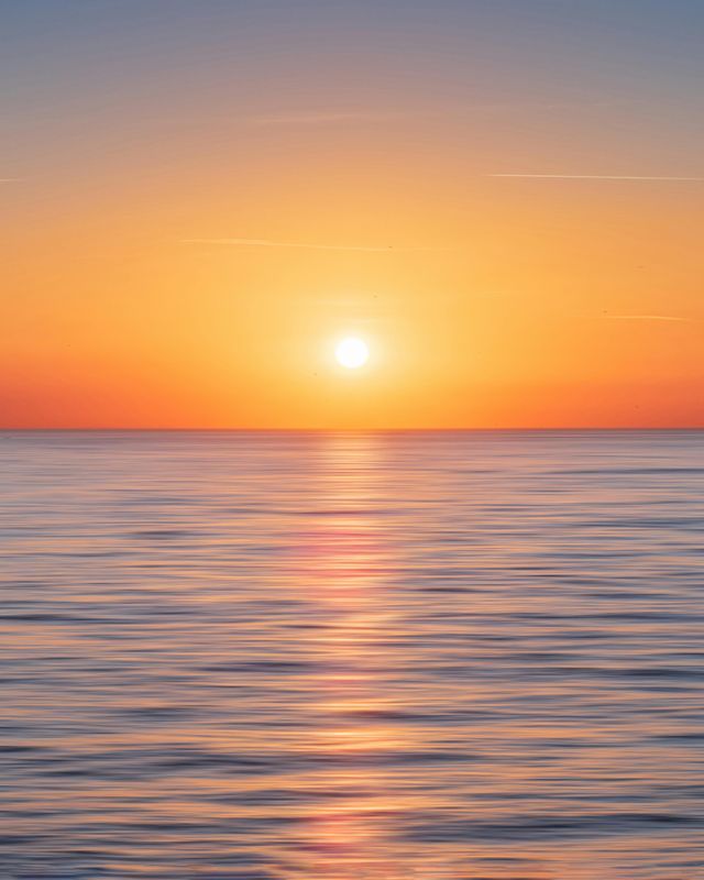 Il sole è basso all'orizzonte, diffondendo un bagliore arancione e giallo su un mare calmo con dolci increspature.