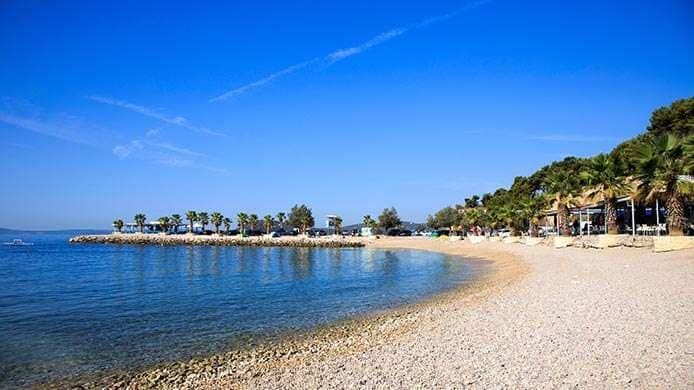 Una spiaggia di ciottoli in una giornata di sole con acqua blu calma in una baia, costeggiata da palme ed edifici.