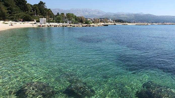 Acqua turchese limpida lambisce una spiaggia di ciottoli, con montagne lontane visibili sotto un cielo azzurro.