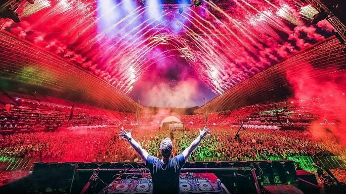 Un DJ con le braccia alzate fronteggia una folla immensa in uno stadio durante un concerto con luci rosse e fuochi d'artificio.